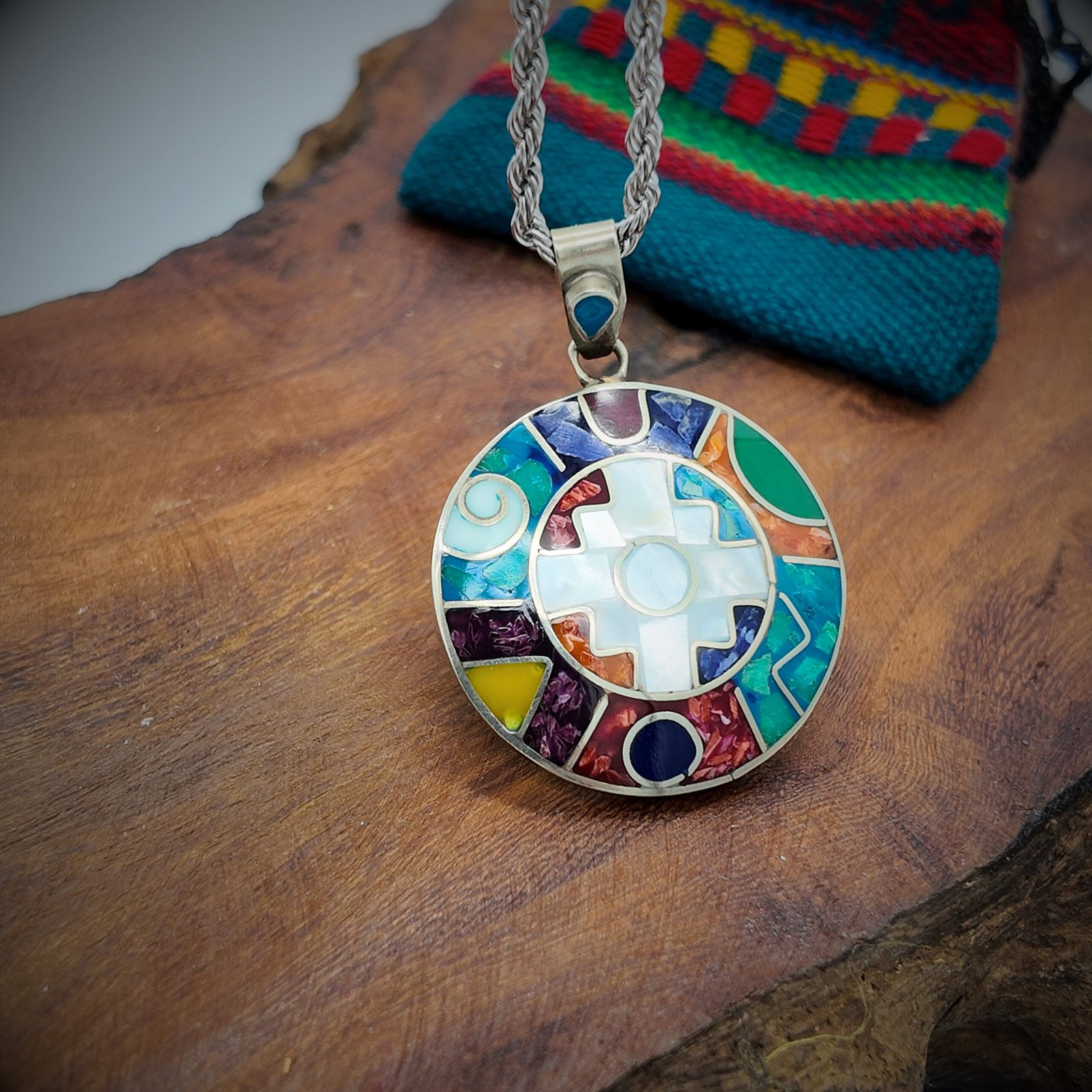 A round disk of artisanal silver with colourful natural stone inlays in a chakana design of the andean shamanic comsology.