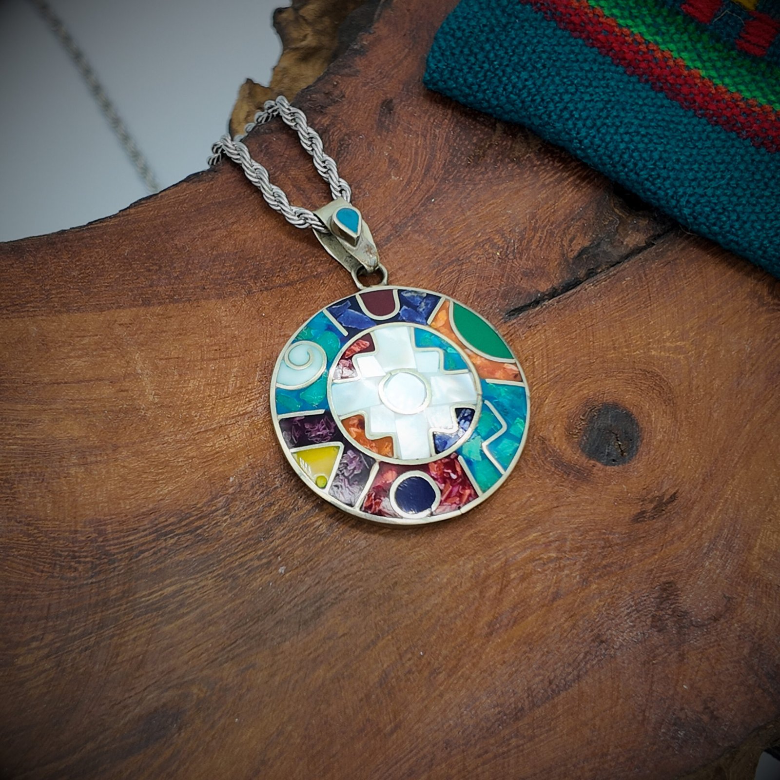 A round disk of artisanal silver with colourful natural stone inlays in a chakana design of the andean shamanic comsology.