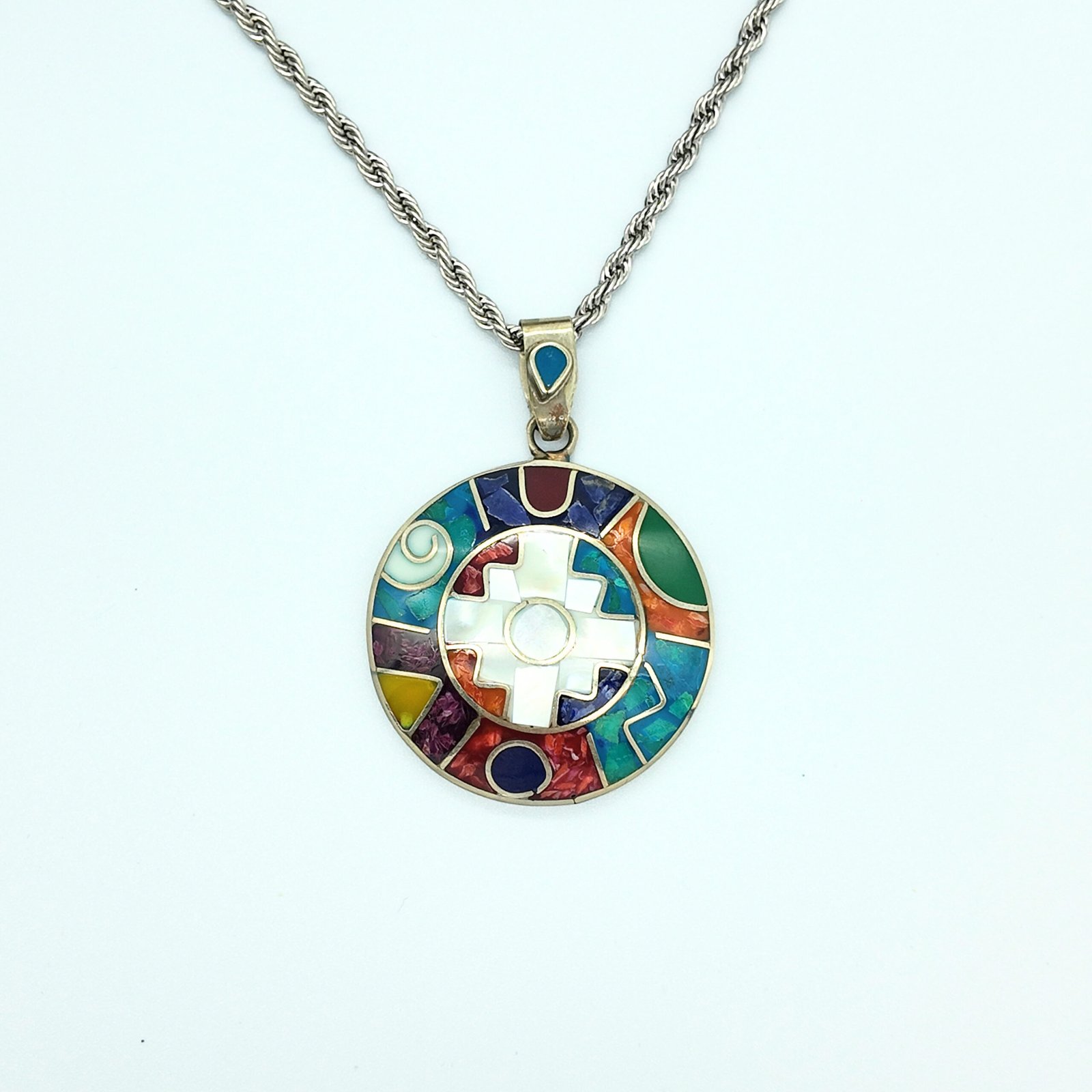 A round disk of artisanal silver with colourful natural stone inlays in a chakana design of the andean shamanic comsology.