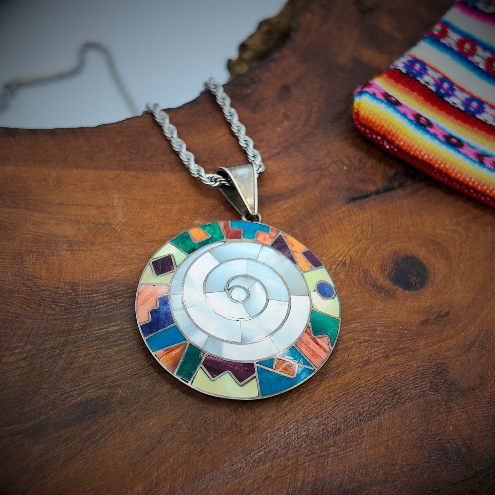 A round disk of artisanal silver with colourful natural stone inlays in a spiral design of the andean shamanic comsology resembling the earth goddess pacha mama cosmic mother