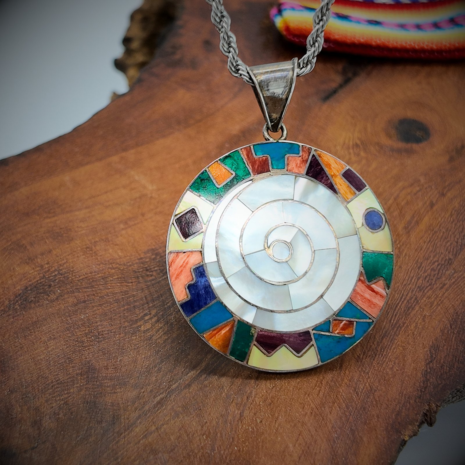 A round disk of artisanal silver with colourful natural stone inlays in a spiral design of the andean shamanic comsology resembling the earth goddess pacha mama cosmic mother