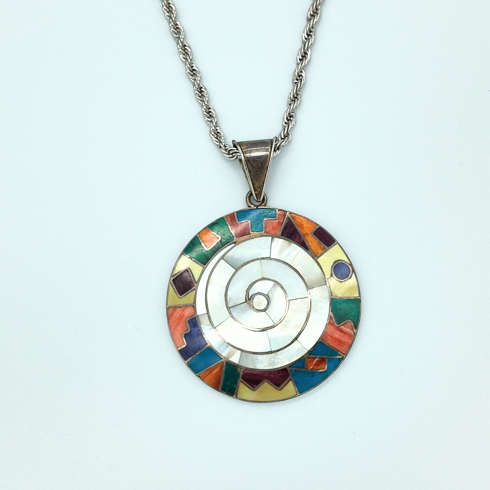 A round disk of artisanal silver with colourful natural stone inlays in a spiral design of the andean shamanic comsology resembling the earth goddess pacha mama cosmic mother